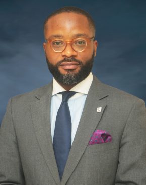 Bespectacled light-skinned man in brown jacket, sky blue and white shirtm dark tie, purple pocket square