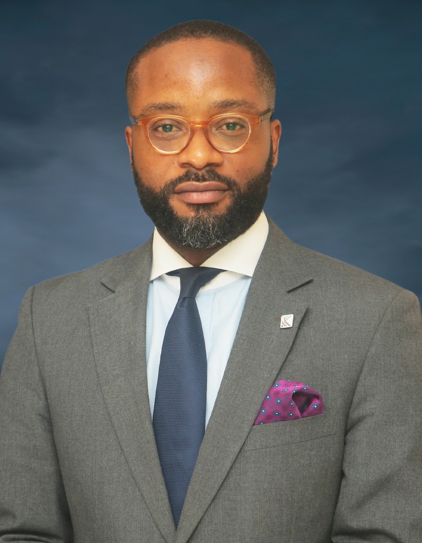 Bespectacled light-skinned man in brown jacket, sky blue and white shirtm dark tie, purple pocket square