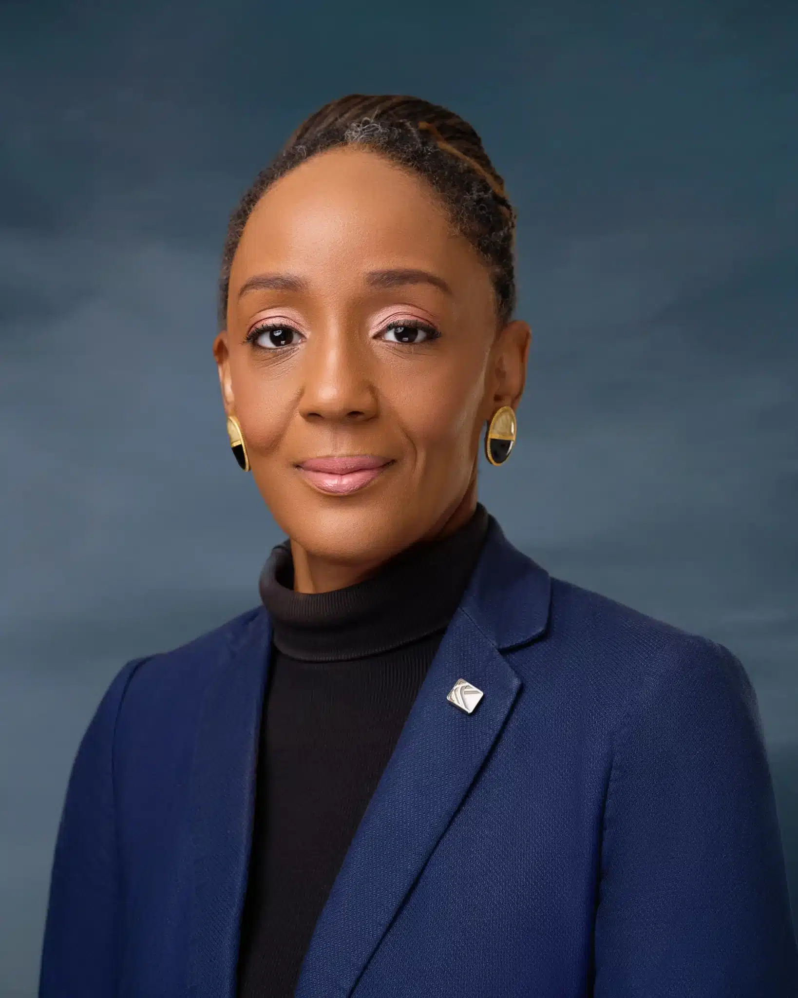 Light-skinned African woman with packed bun, blue jacket, black turtle neck and big gold and black earrings