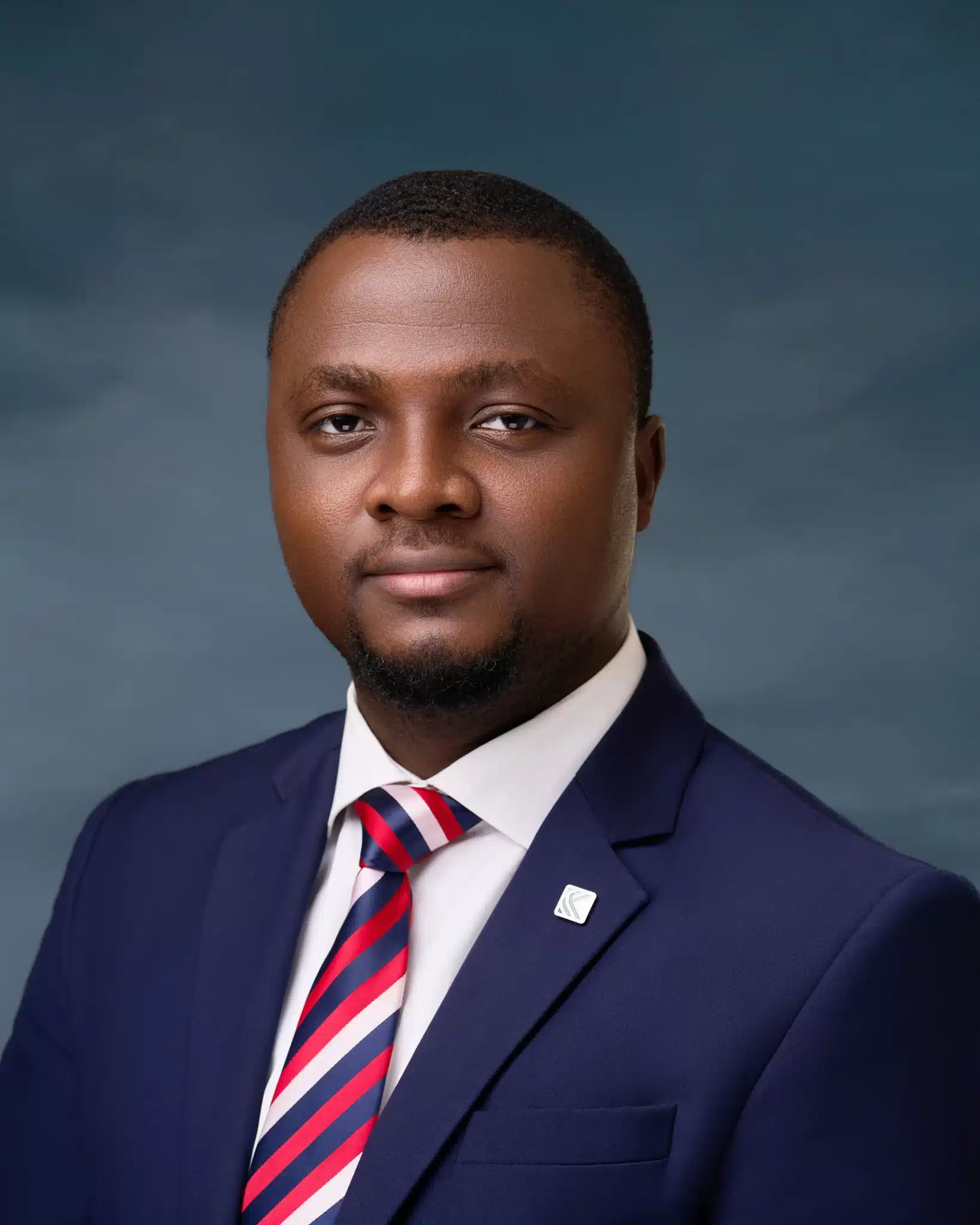 Dark-skinned man in a blue jacket and white shirt, sporting a multicoloured tie and a lapel pin