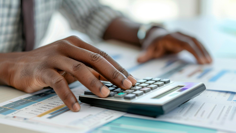 AI-generated image of the hands of a black man with rolled sleeves and wristwatch using a calculator on a table with spreadsheets